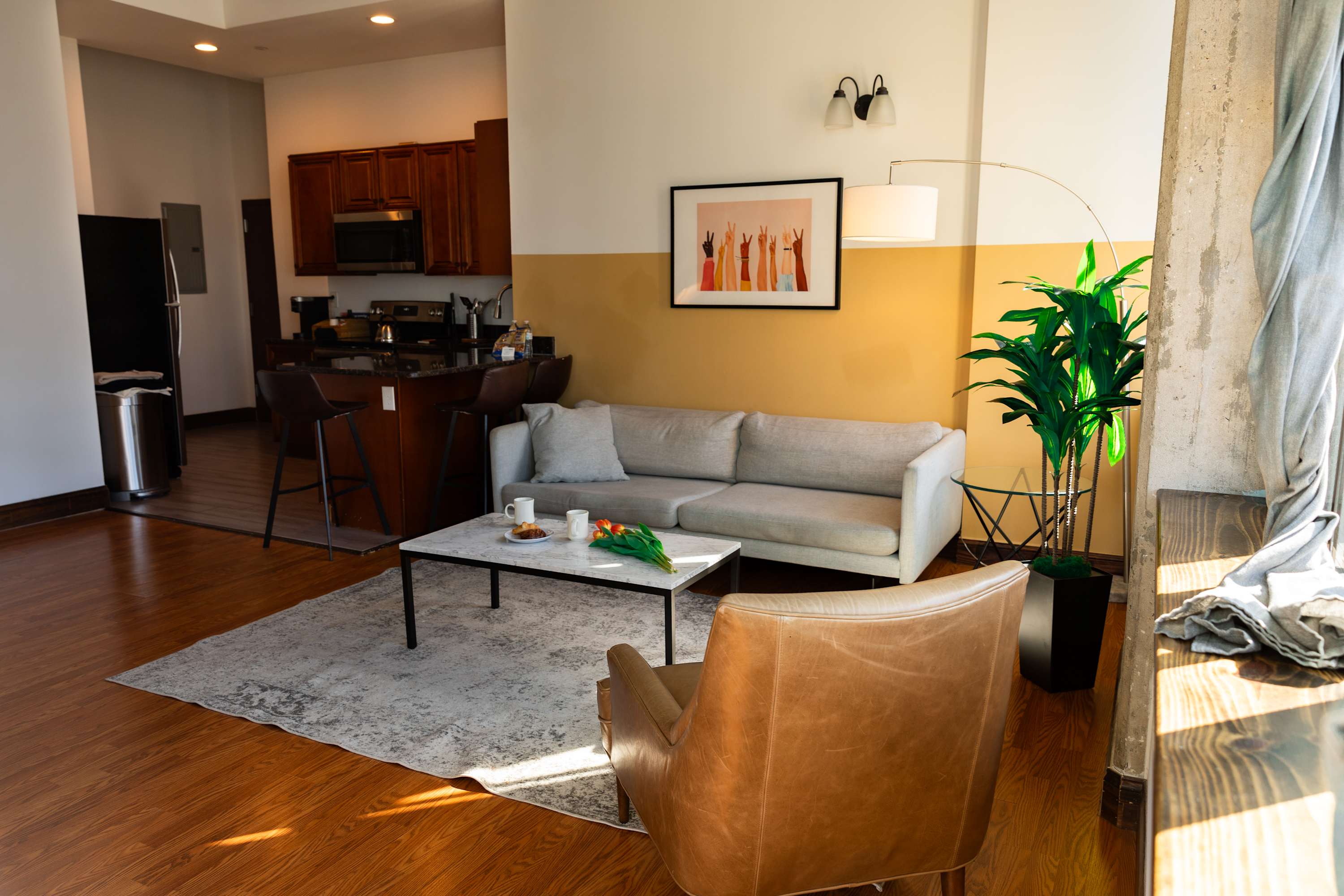 Spacious living room with a light grey sofa, a brown leather armchair, and a marble coffee table on a patterned rug. The room features two-toned walls, warm hardwood floors, and an open view towards the kitchen area with wooden cabinets and a breakfast bar.