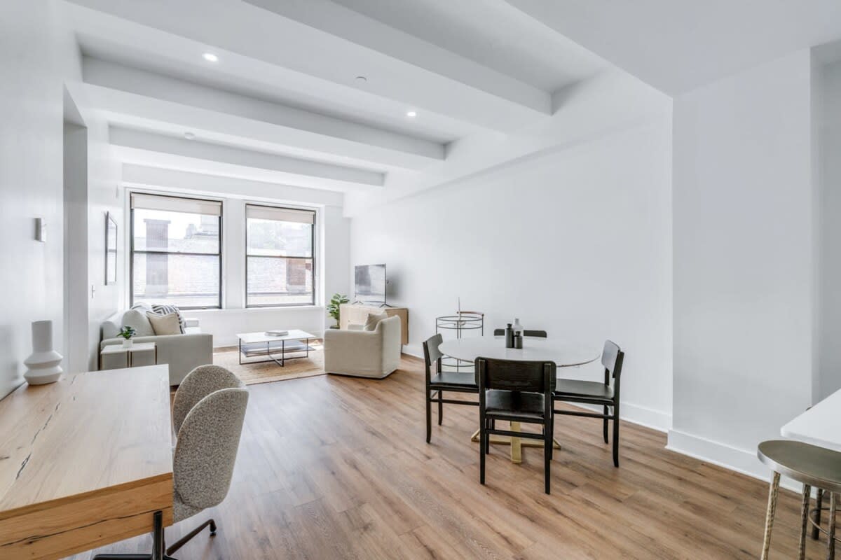 Expansive open-concept living space with light hardwood floors, featuring a living area with a sofa and armchair, a dining area with a round table and black chairs, and a small desk nook. Large windows illuminate the room.
