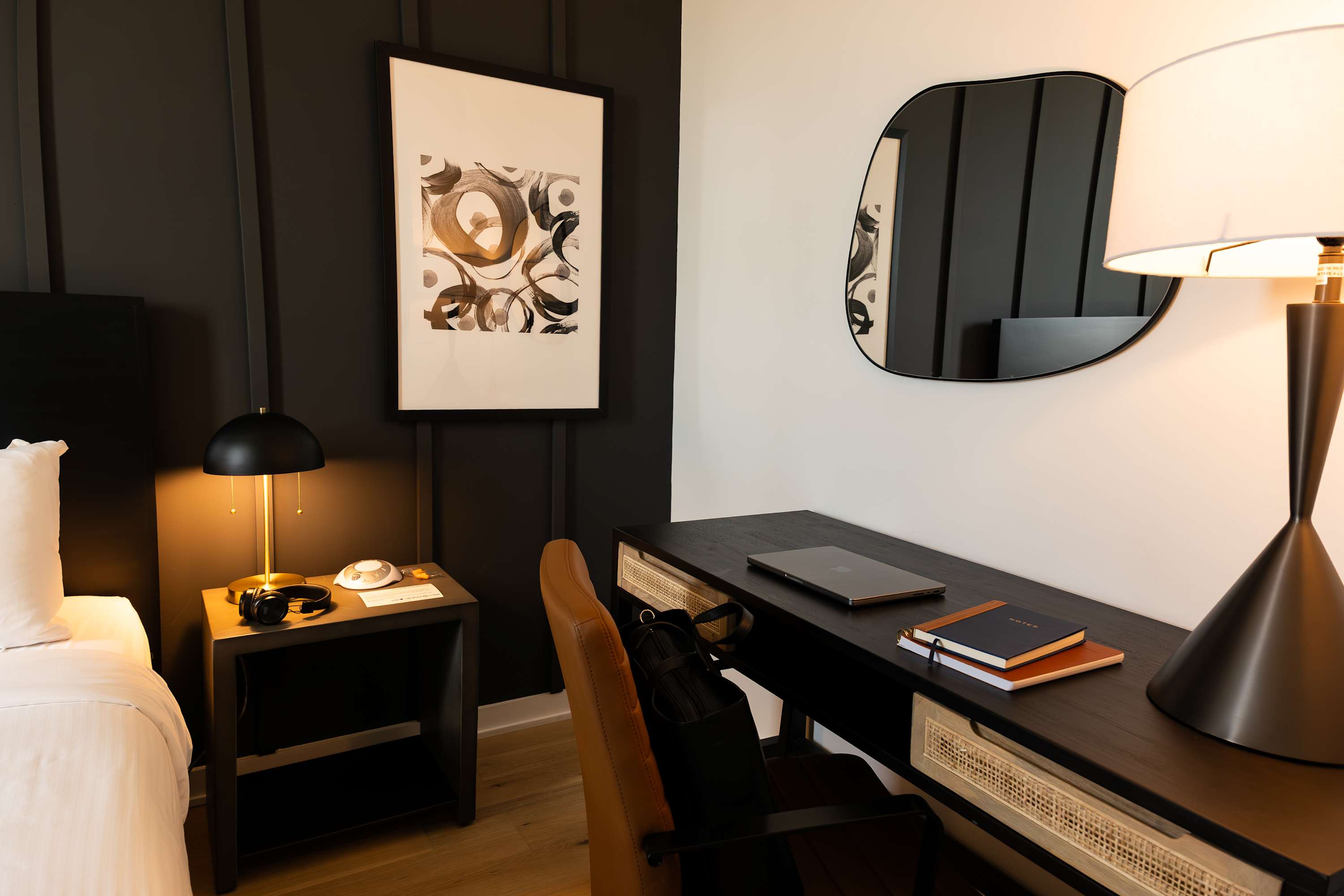 A functional workspace within a bedroom, featuring a dark wooden desk with rattan drawer fronts, a brown leather office chair, and a large black-framed mirror. A laptop and notebooks are neatly arranged on the desk, with a bedside lamp visible on the left.