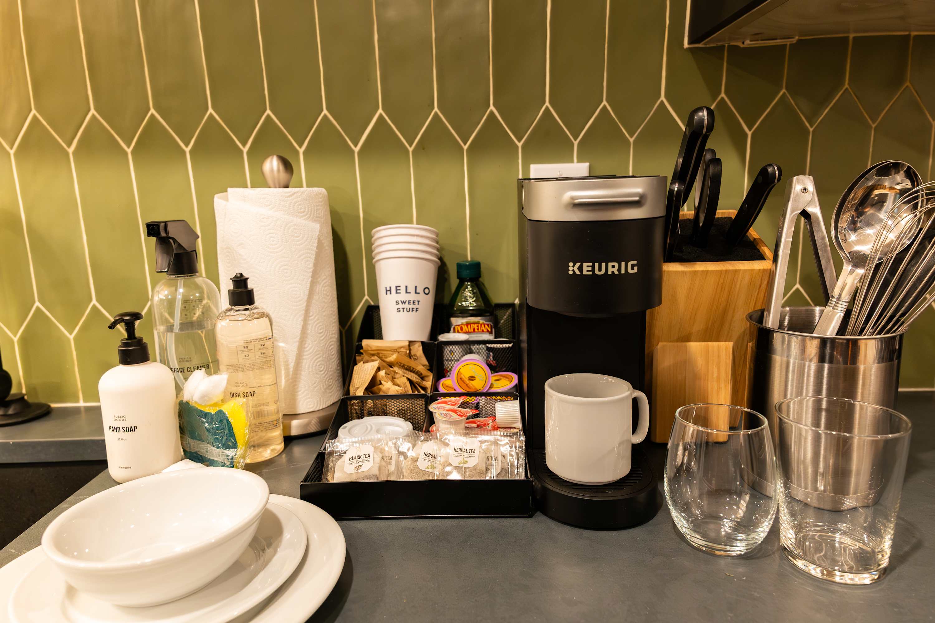A well-stocked kitchen counter with a green geometric tile backsplash features a Keurig coffee maker, various tea and coffee supplies, paper towels, and cleaning supplies, alongside white dishes and glassware.