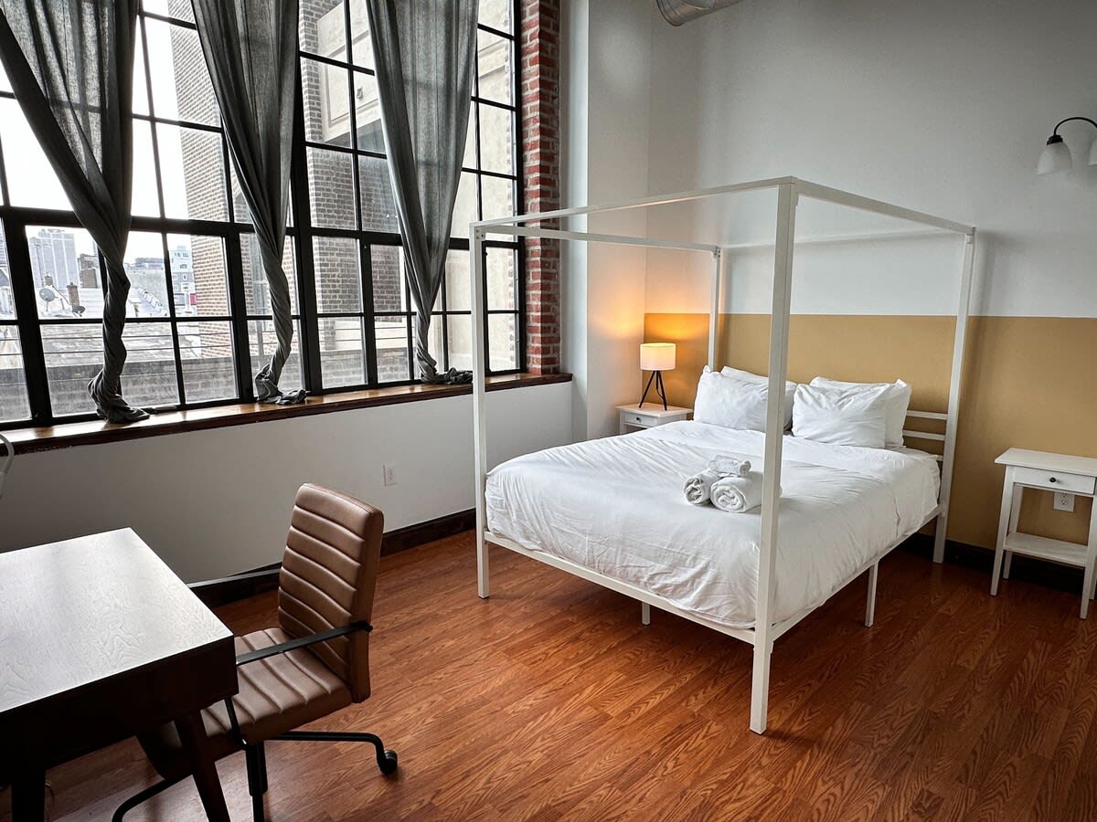 Spacious bedroom featuring a white four-poster bed with white linens, a brown leather office chair, and a dark wooden desk, illuminated by large industrial-style windows with dark gray curtains and a view of the city.