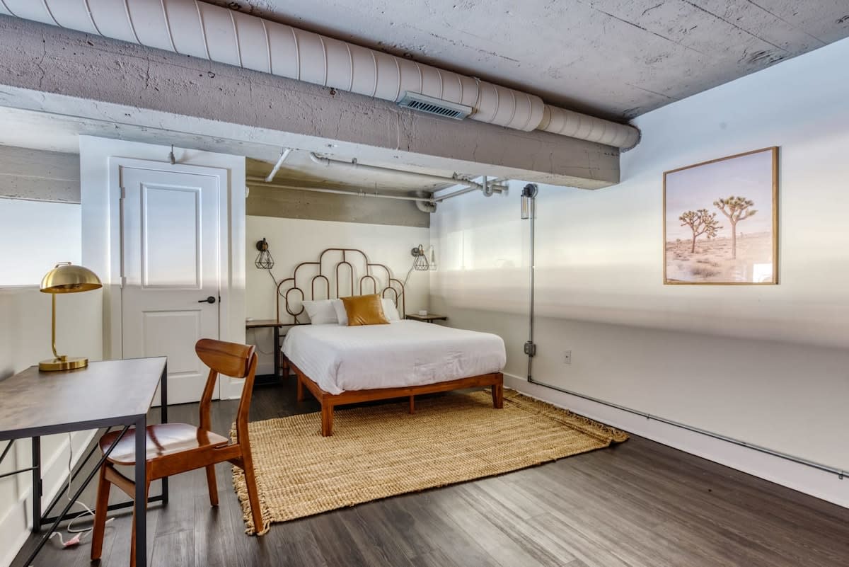 A cozy loft bedroom with a queen-sized bed featuring a decorative metal headboard and a mustard yellow pillow, set on a natural fiber rug. A wooden desk with a gold lamp and chair is to the left, and exposed concrete ceiling with ductwork adds an industrial touch.