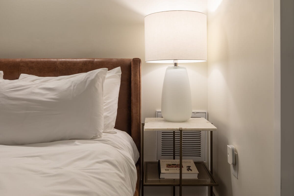 A close-up of a white-shaded lamp on a small marble-topped nightstand, positioned next to a bed with crisp white linens and a brown upholstered headboard. A book rests on the lower shelf of the nightstand.