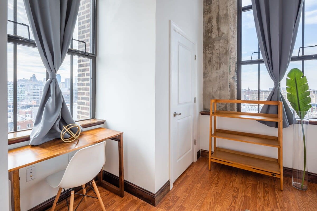 A dedicated office nook featuring a simple wooden desk and a white modern chair, positioned beside a tall industrial window with a city skyline view. A small wooden bookshelf and a decorative plant add warmth to the space.