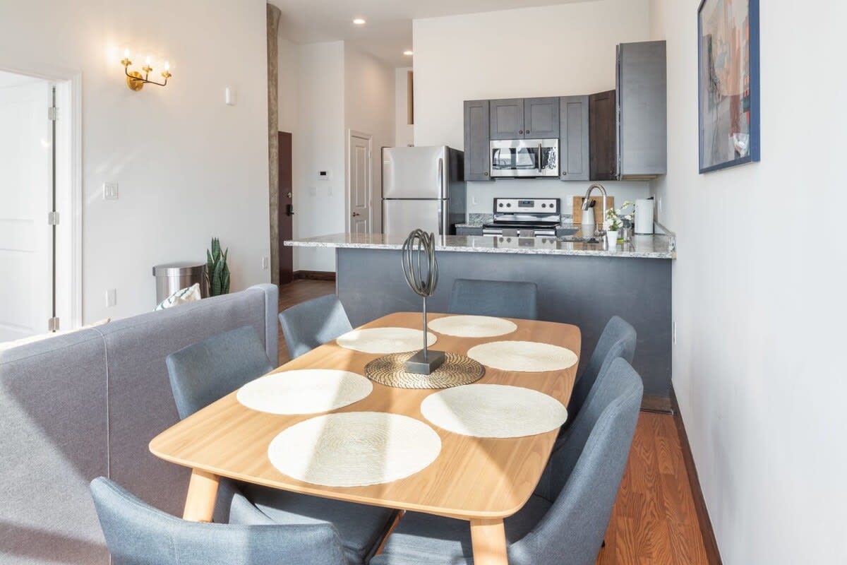 Open-concept dining area with a light wood rectangular table and six grey upholstered chairs, leading into a modern kitchen with dark cabinetry and stainless steel appliances. The space features hardwood floors and white walls.