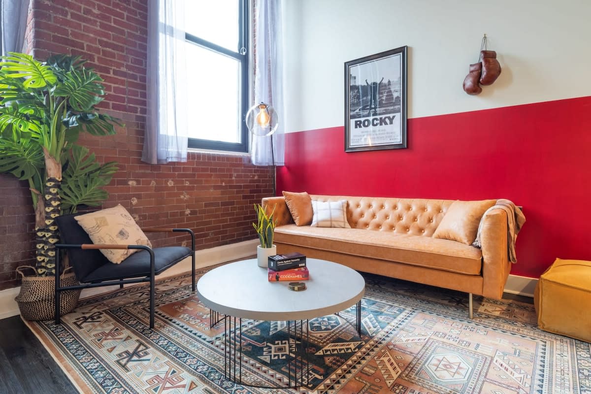 Inviting living room featuring a tan tufted sofa, a round concrete coffee table, and a vibrant patterned area rug. Exposed brick and a bold red accent wall are decorated with a "Rocky" poster and boxing gloves.