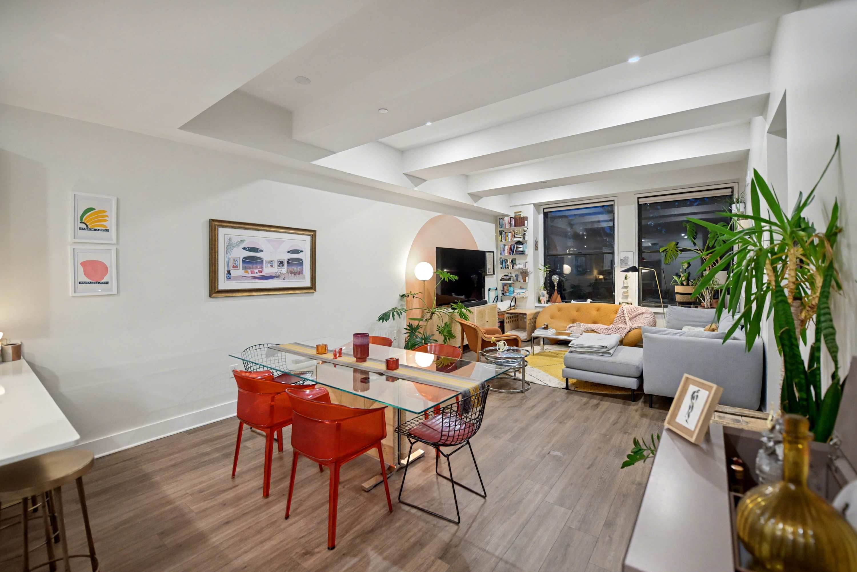An open-concept living space combines a dining area with a glass table and red chairs, a white kitchen, and a living room with a gray sectional and orange sofa. The space is unified by hardwood floors and a peach accent wall.