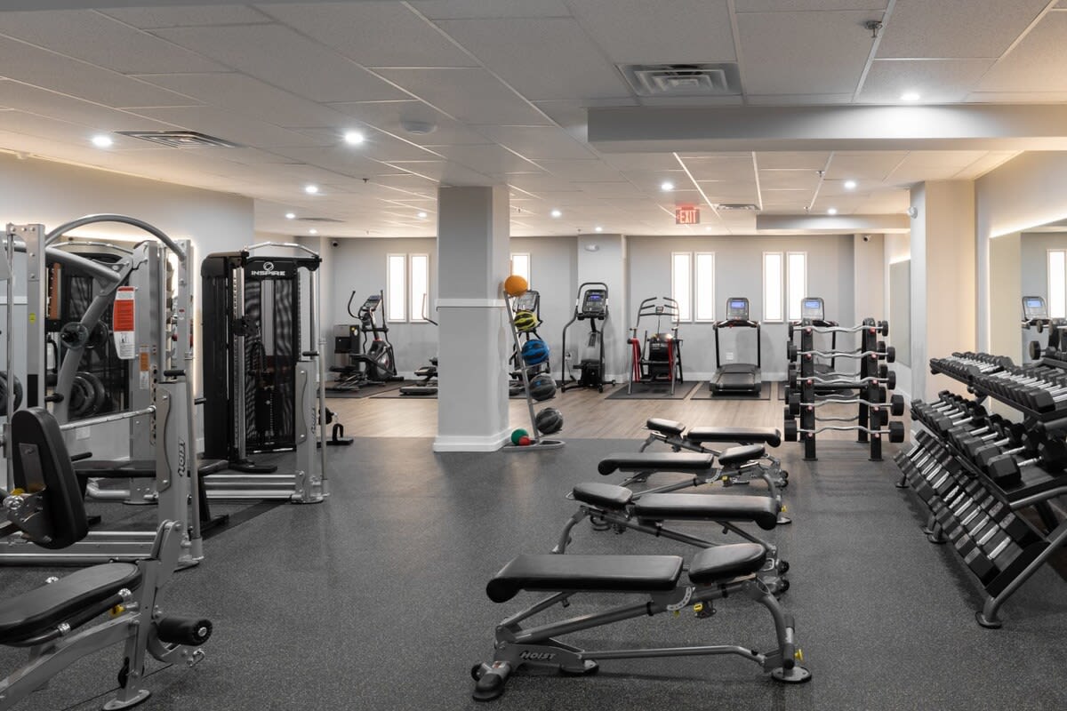 Spacious communal gym with various exercise equipment including treadmills, ellipticals, weight machines, and free weights. The floor is covered with black rubber mats, and large mirrors line one wall, enhancing the open feel.