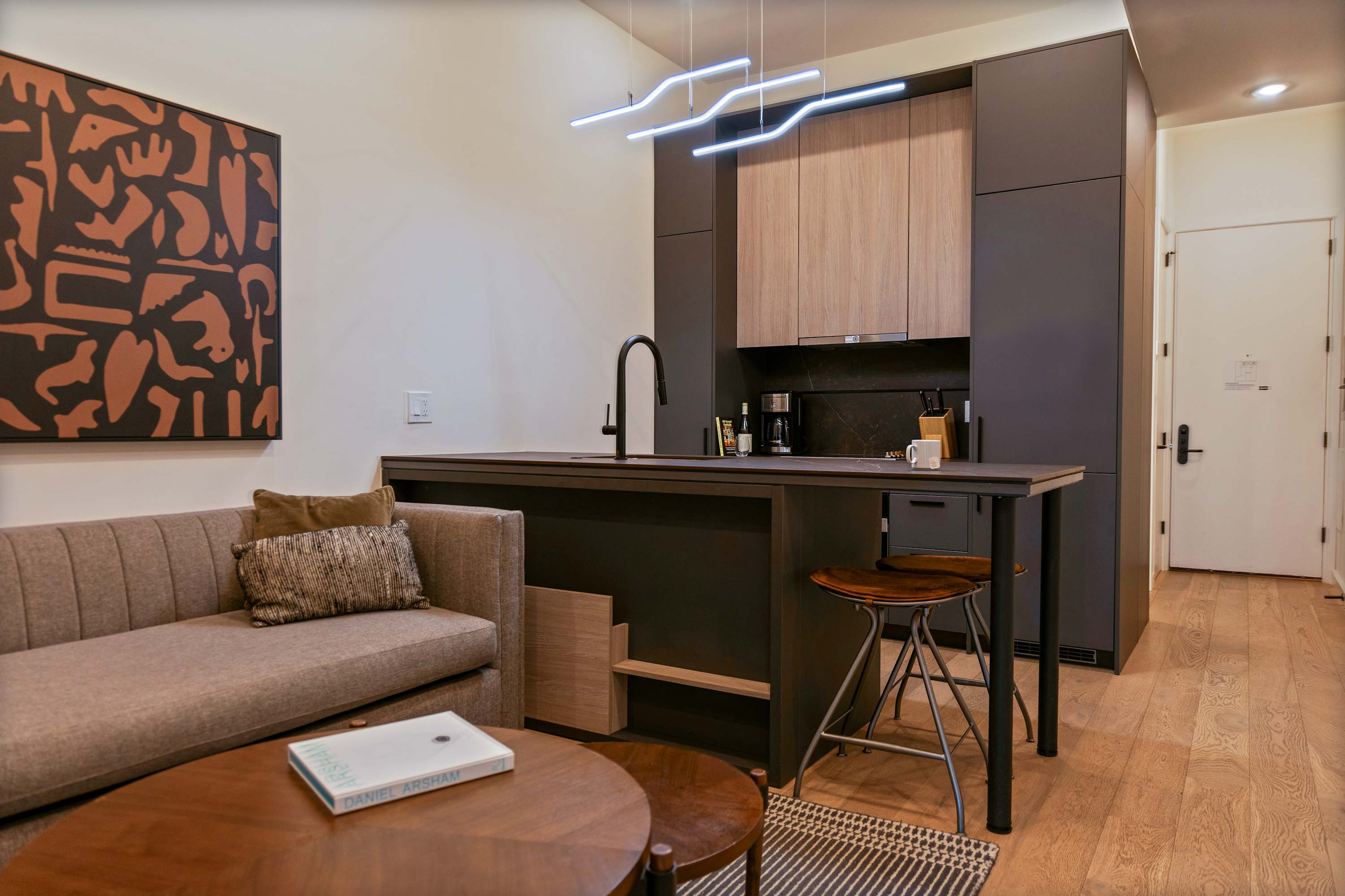 Contemporary open-concept living area featuring a neutral-toned sofa, round wooden coffee tables, and a sleek kitchen island with a black faucet and bar stools. Modern linear pendant lighting illuminates the space, which has light wooden flooring.