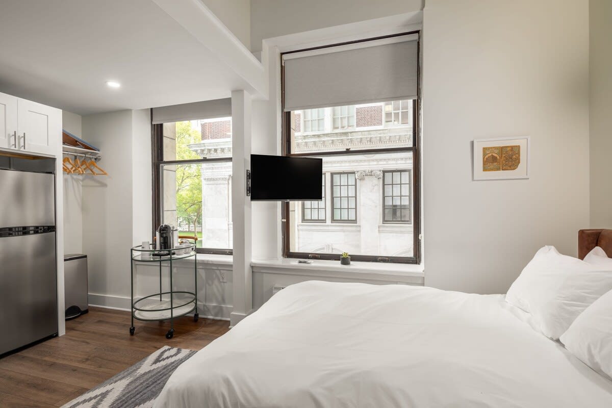 An elevated view of the studio apartment, showcasing a queen bed with white linens, large windows with room-darkening shades and city views, and a partial view of the kitchenette on the left. A flat-screen TV is mounted on the wall near the windows.