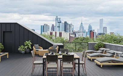 Expansive rooftop deck with a stunning city skyline view, featuring a dining area with a large table and chairs, multiple lounge chairs, and a separate seating area with wicker furniture. The deck has dark flooring and lush greenery.
