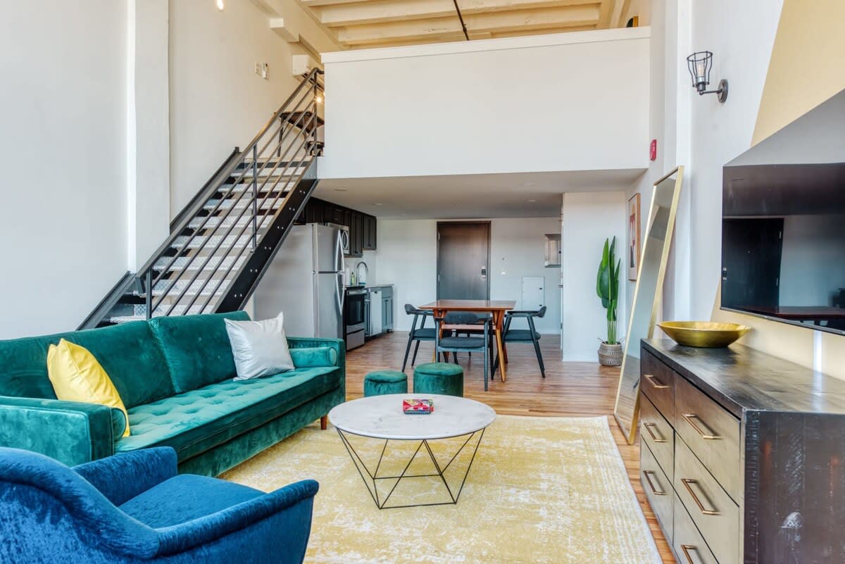 Open-concept living space with a teal tufted sofa, a white and gold coffee table, and a metal staircase leading to an upper loft. The view extends to a dining area and a modern kitchen with dark cabinetry.
