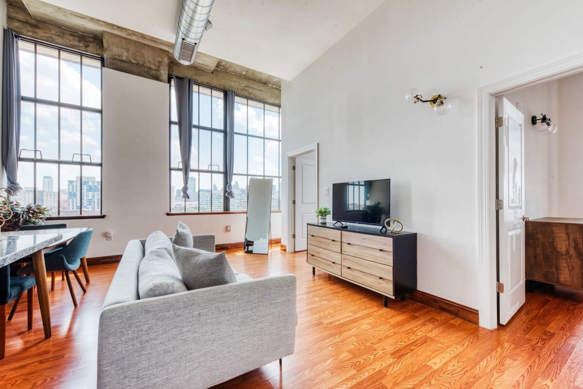 A bright living room featuring a comfortable light gray sofa facing a modern wooden dresser topped with a television. Expansive industrial windows provide a stunning city panorama, and a full-length mirror leans against the wall.