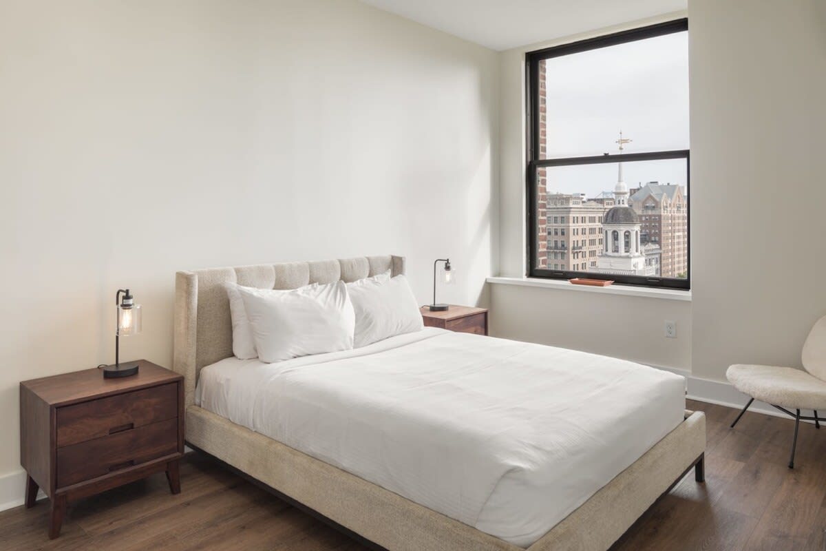Cozy bedroom with a queen-sized upholstered bed, two dark wood nightstands with modern lamps, and a large window overlooking city buildings.