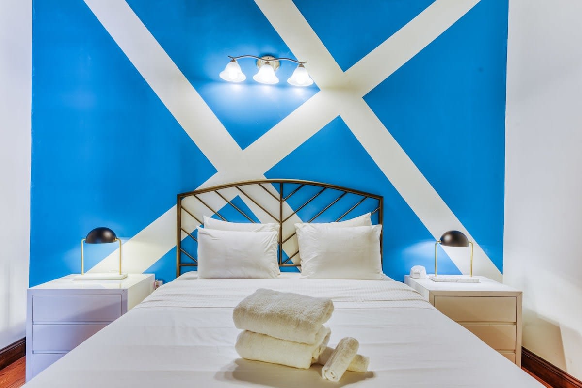Bedroom featuring a queen bed with a black metal frame and white bedding, flanked by two white nightstands with black lamps. A striking blue and white geometric pattern is painted on the accent wall behind the bed.