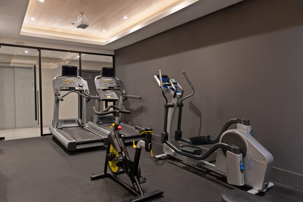 Well-equipped fitness room with dark gray walls and a black rubber floor, featuring two treadmills, an elliptical machine, and a stationary bike. A glass partition separates the gym from another area, with subtle overhead lighting.