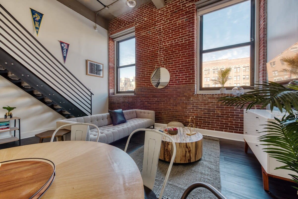 Modern loft living space with exposed brick walls and large windows, featuring a light gray tufted sofa, a round wooden coffee table, and a round dining table with light blue and black chairs. A black metal staircase leads to an upper level.