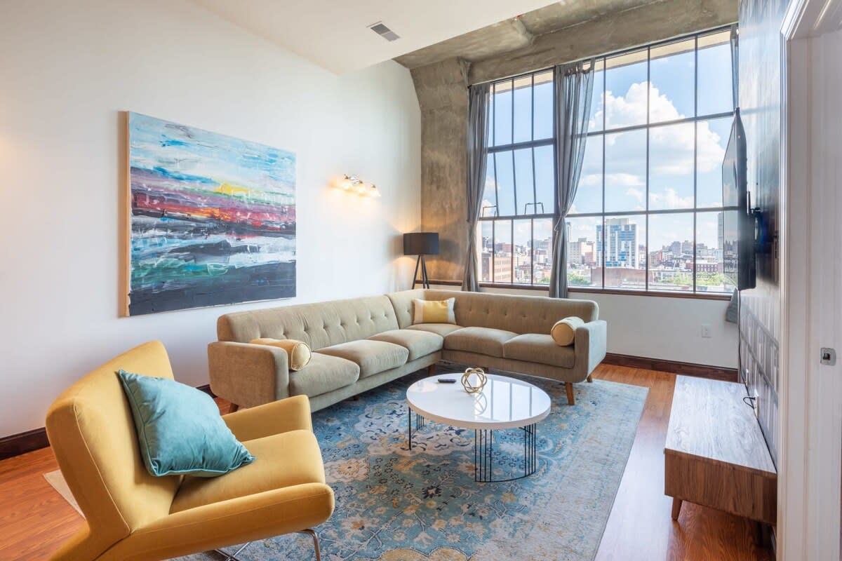 Spacious living room with a light-colored sectional sofa, a yellow accent chair, a white round coffee table, and a large abstract painting, all set on a patterned area rug.