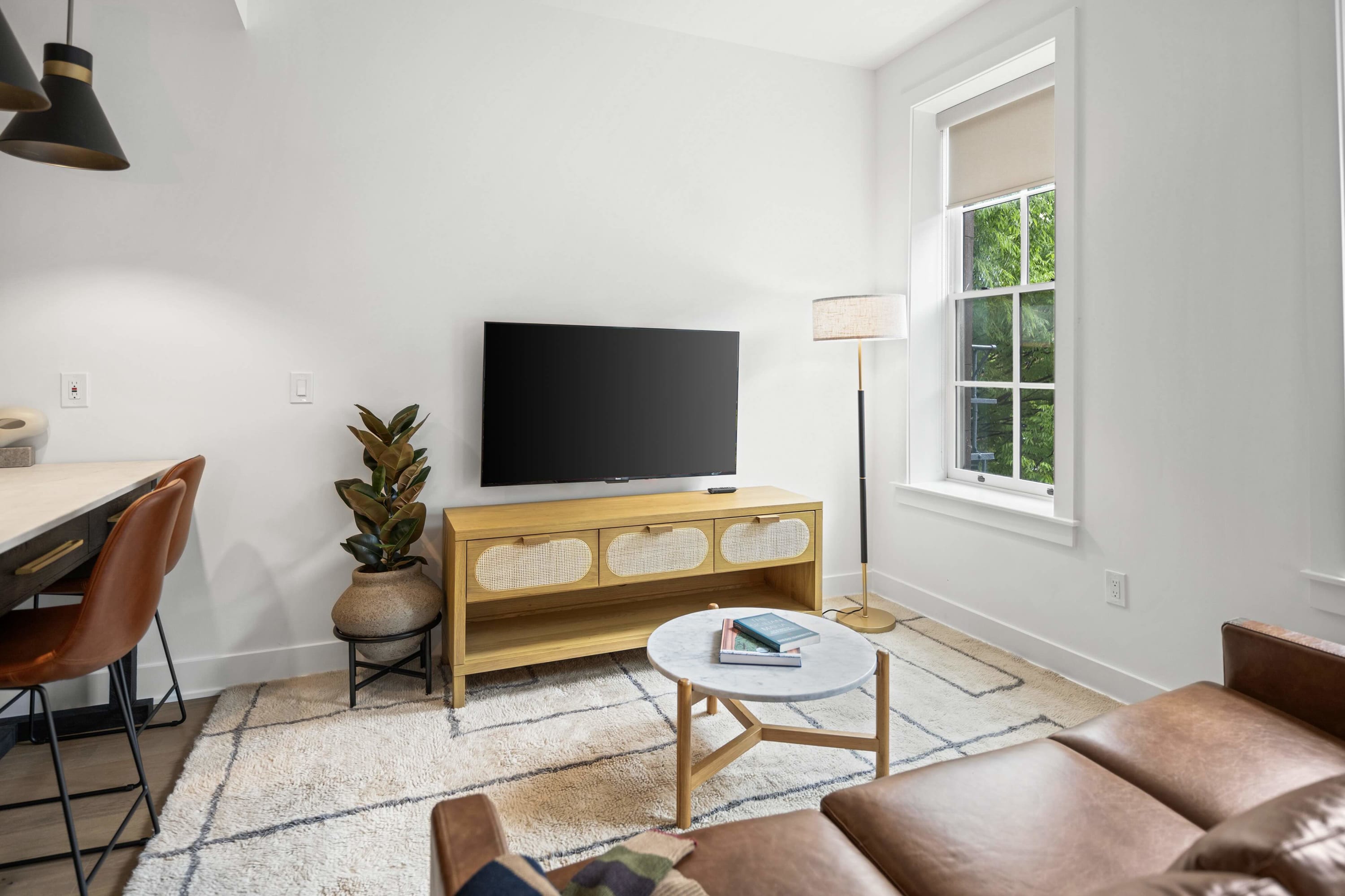 Cozy living room featuring a brown leather sofa, a large flat-screen TV on a light wood and rattan console, and a round marble coffee table. The room is bright with white walls, a patterned area rug, and a tall floor lamp.