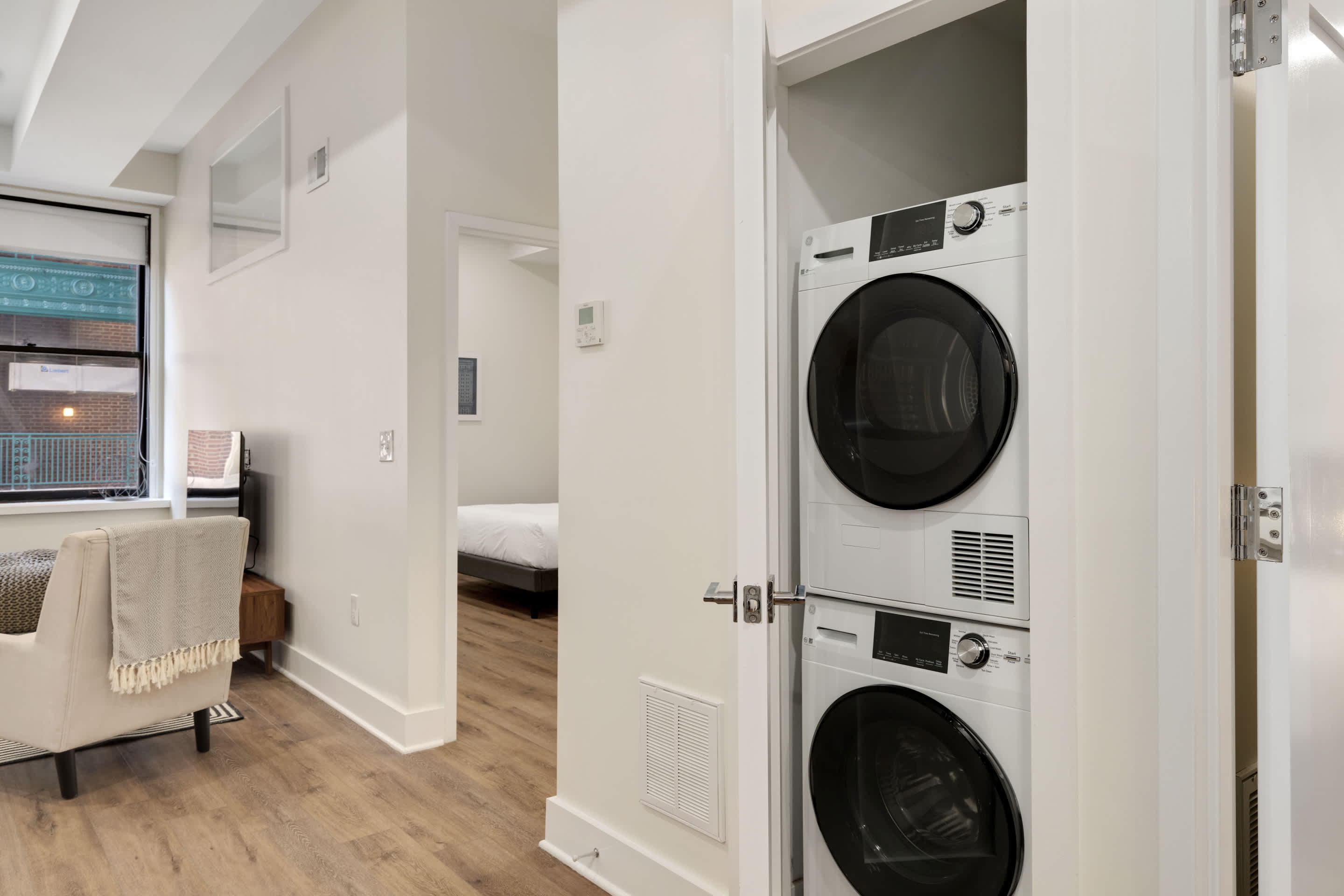 A stacked white washer and dryer unit is neatly installed within a closet, with a closed door visible to the left. The view extends into a hallway with light hardwood floors, offering a glimpse of a bedroom in the background.