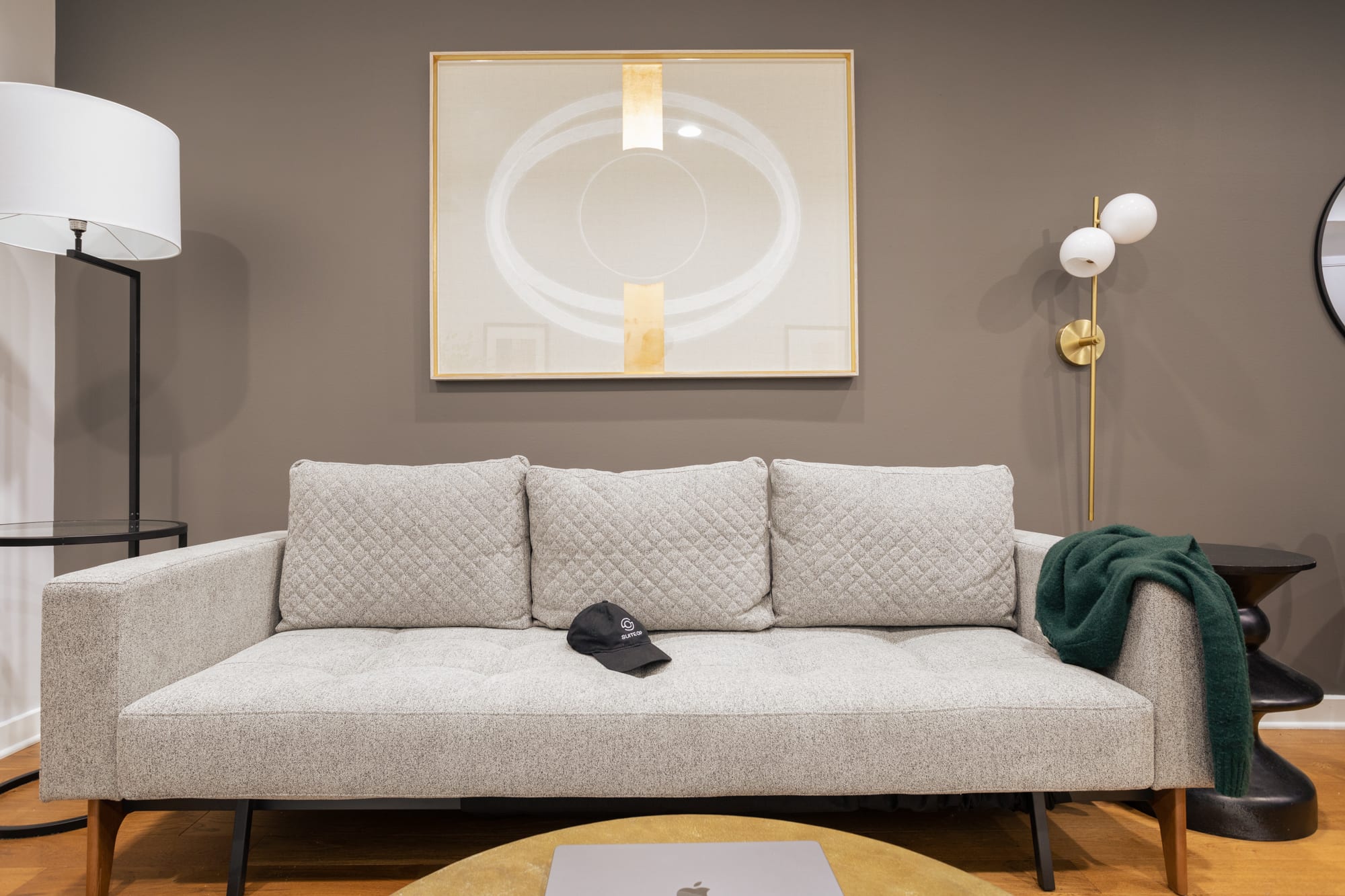 A stylish living room showcasing a light grey quilted sofa with a green throw blanket and a black baseball cap. Above the sofa, a large framed abstract artwork with gold accents adorns the dark grey wall.
