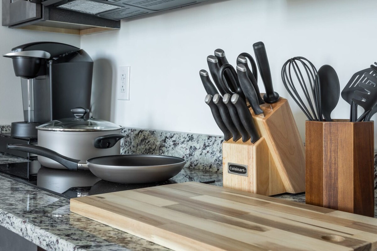 A detailed view of a kitchen countertop, showcasing a coffee maker, a pot, a frying pan, a wooden knife block filled with knives, and a utensil holder. The granite countertop is complemented by a wooden cutting board.