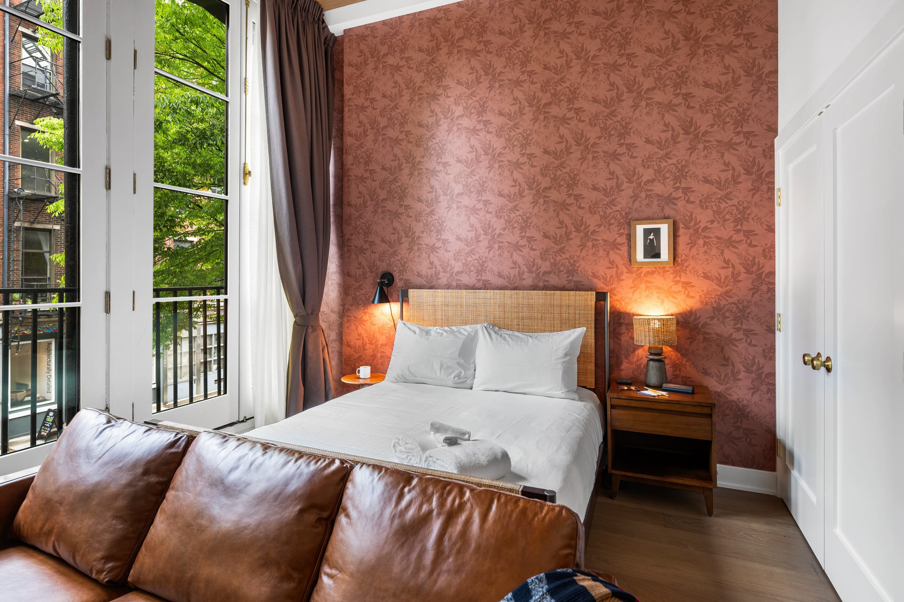 A wide shot of the studio apartment, featuring a queen bed with a rattan headboard against a floral accent wall, and a brown leather sofa in the foreground. French doors open to a Juliet balcony, revealing a vibrant city view with green trees.