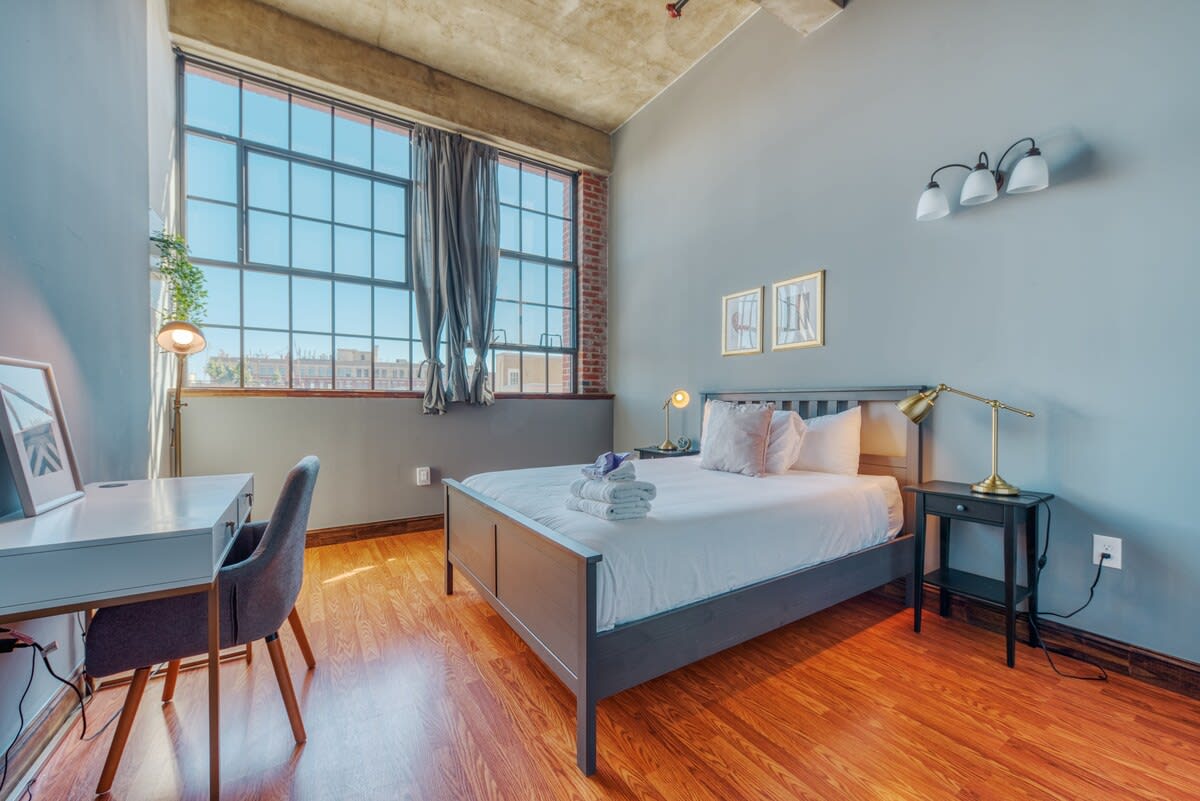 A bright bedroom features a dark gray bed with white bedding, a light gray desk with a matching chair, and large industrial-style windows with brick accents, providing ample natural light.