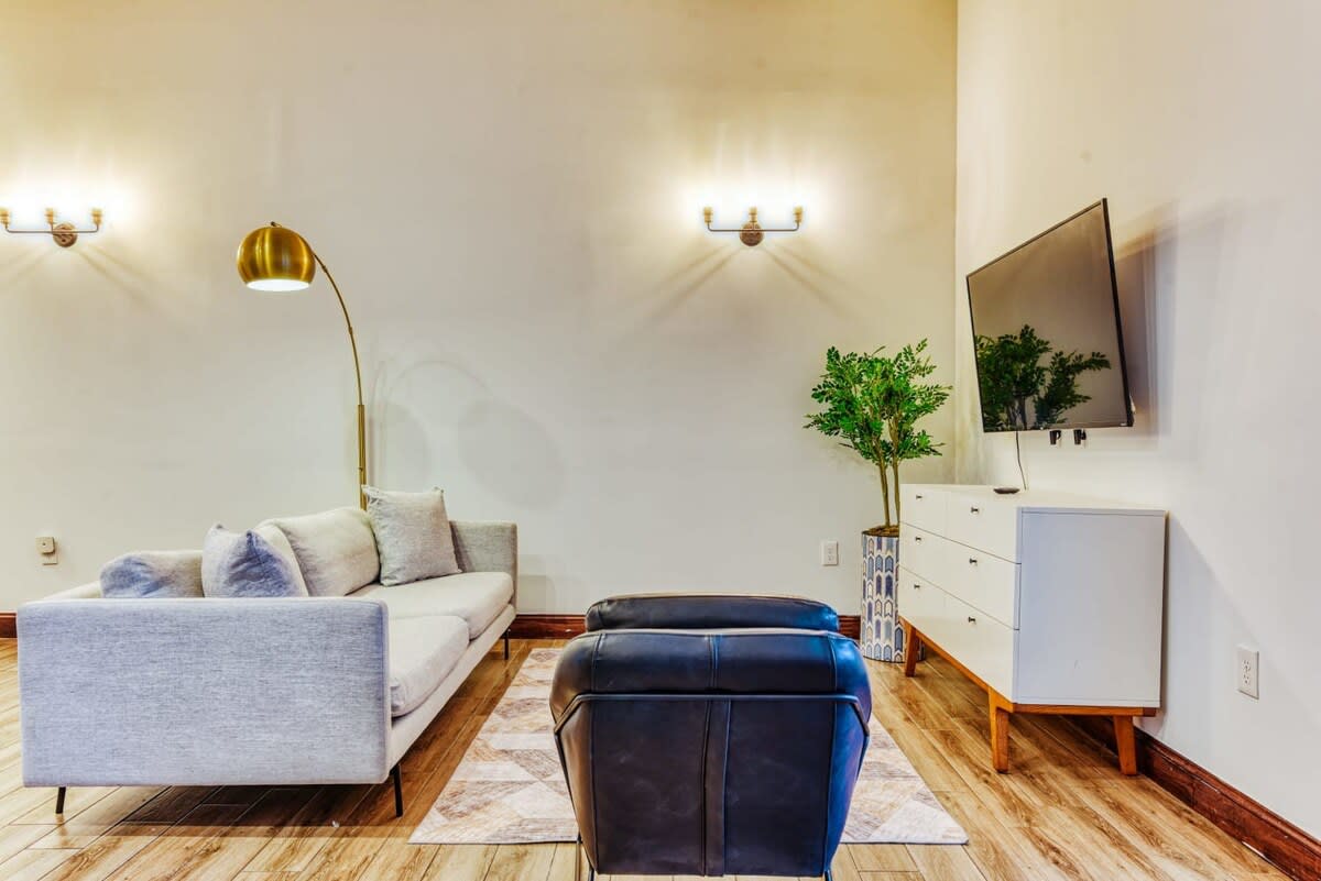 A modern living room with light wood-look flooring and white walls, featuring a light grey sofa with black legs and a dark leather armchair on a geometric patterned rug. A large wall-mounted TV is positioned above a white and wood dresser, with a tall brass floor lamp providing additional lighting.