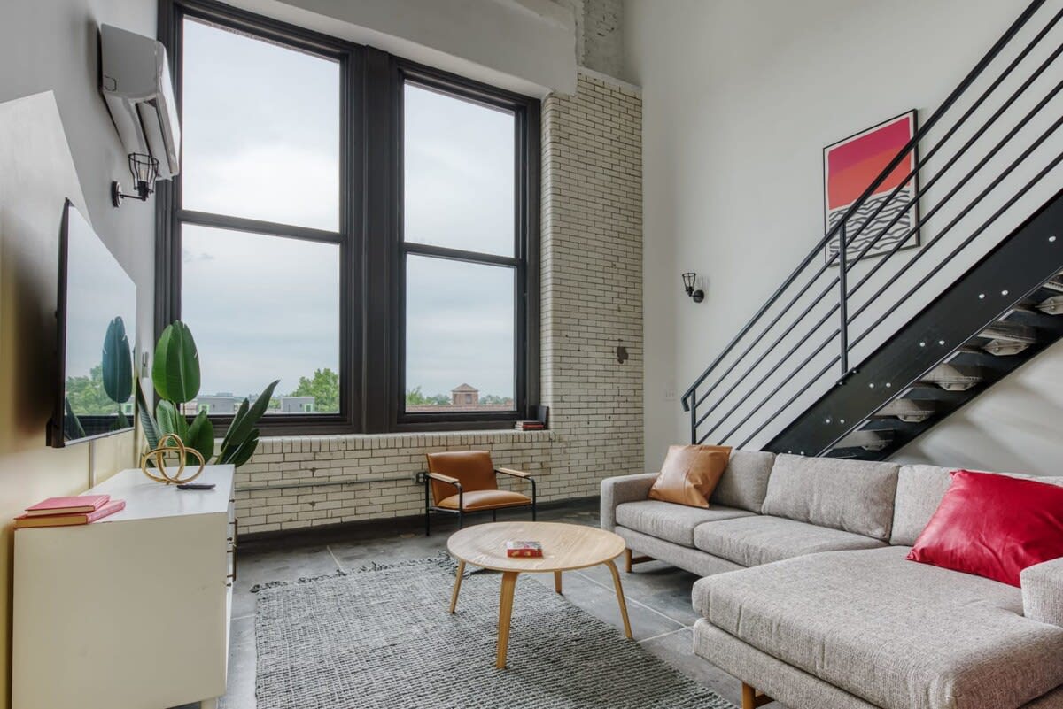Airy living room featuring a light gray sectional sofa, a round wooden coffee table, and a brown leather accent chair, all resting on a gray area rug over a concrete floor. Tall, dark-framed windows provide ample natural light and a view.