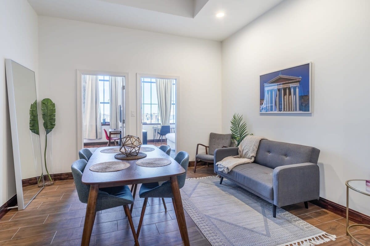 A modern open-concept living and dining area with white walls and wood-look tile flooring. The dining table is set for four, and the living space includes a gray sofa and armchair, with large windows visible through doorways.