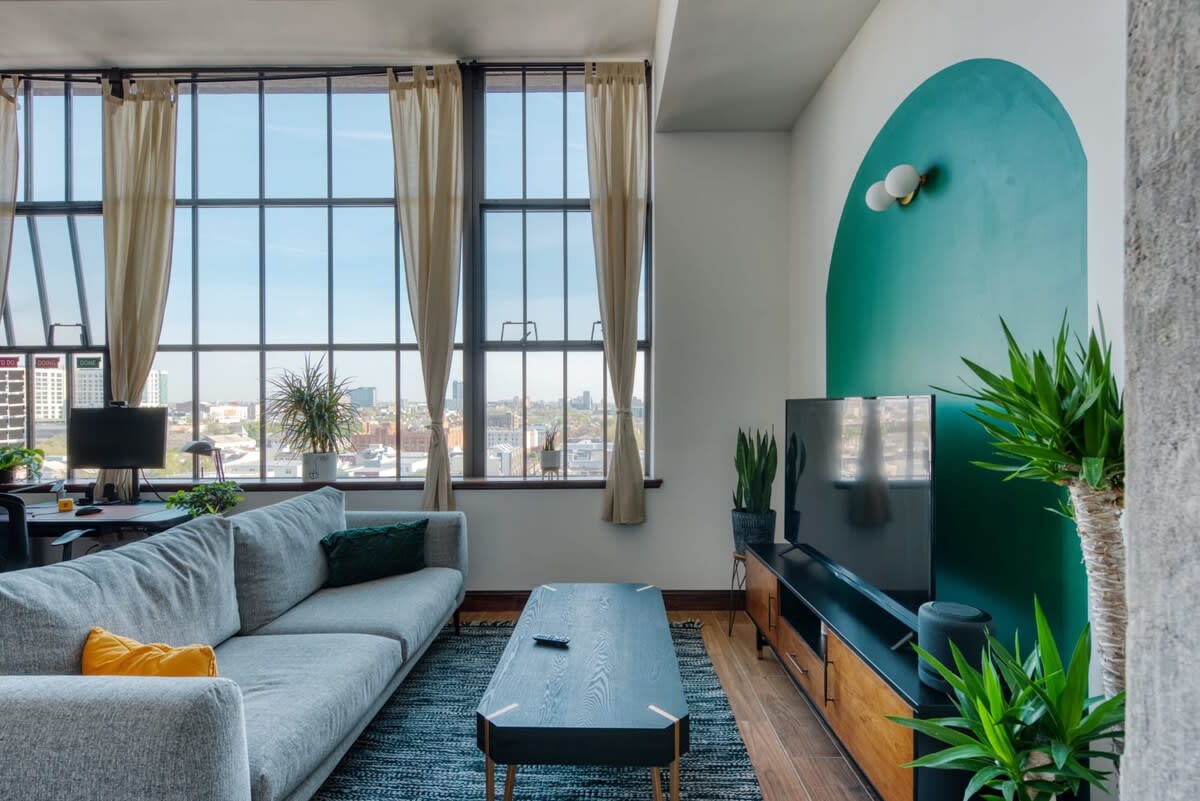 A wide shot of the modern living room shows a light gray sectional sofa, a black rectangular coffee table on a patterned rug, and a large TV console. Tall industrial-style windows with light curtains overlook a city view, and a desk with a computer is visible to the left.