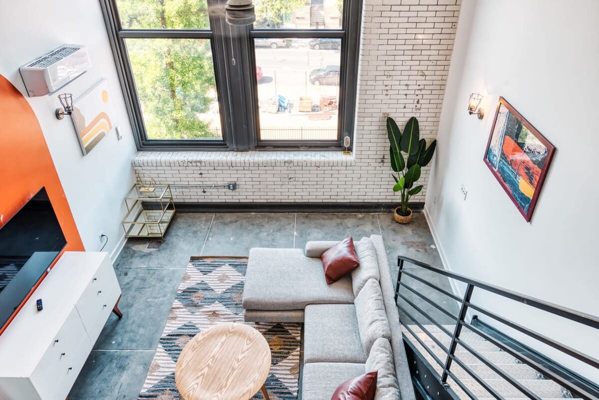 Aerial view of a stylish loft living room featuring a light gray L-shaped sectional sofa with red accent pillows, a round wooden coffee table, and a geometric patterned rug. A white media console with a TV is against an orange accent wall, with large windows and a white brick wall visible.