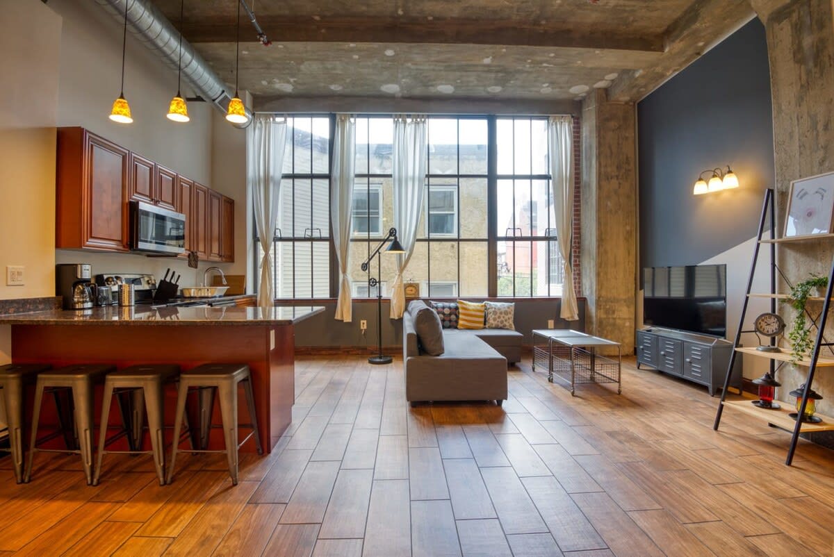 An open-concept living space with a grey sectional sofa facing a large industrial window, adjacent to a kitchen island with bar stools and wooden cabinetry, all under a concrete ceiling with exposed pipes.