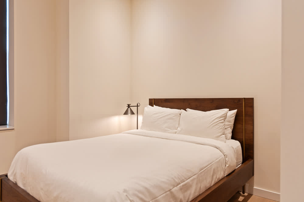 Cozy bedroom with a wooden bed frame and headboard, crisp white bedding, and a small black task lamp on a bedside table, set against light-colored walls.