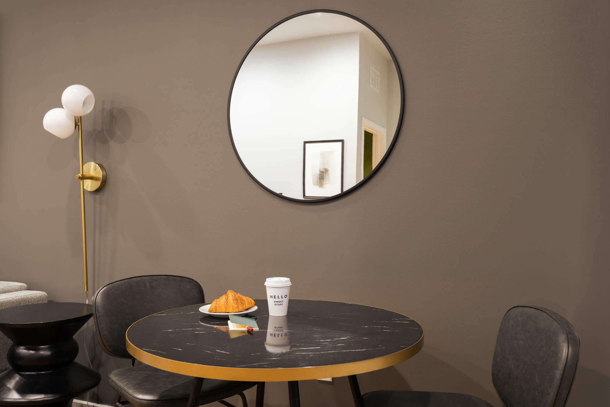 A compact dining area featuring a round black marble-top table with gold accents and two black leather chairs. A large circular mirror hangs on the dark grey wall, and a modern floor lamp provides ambient lighting.