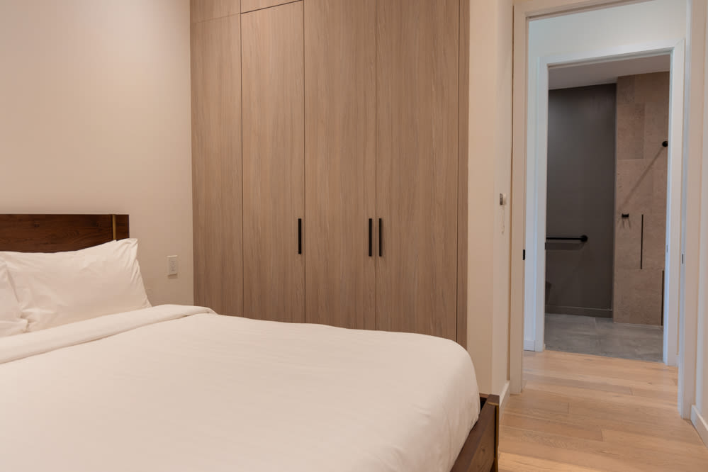 View of the bedroom showing a wooden bed with white bedding, facing a wall with light wood built-in wardrobes with black handles, and an open doorway leading to a bathroom.