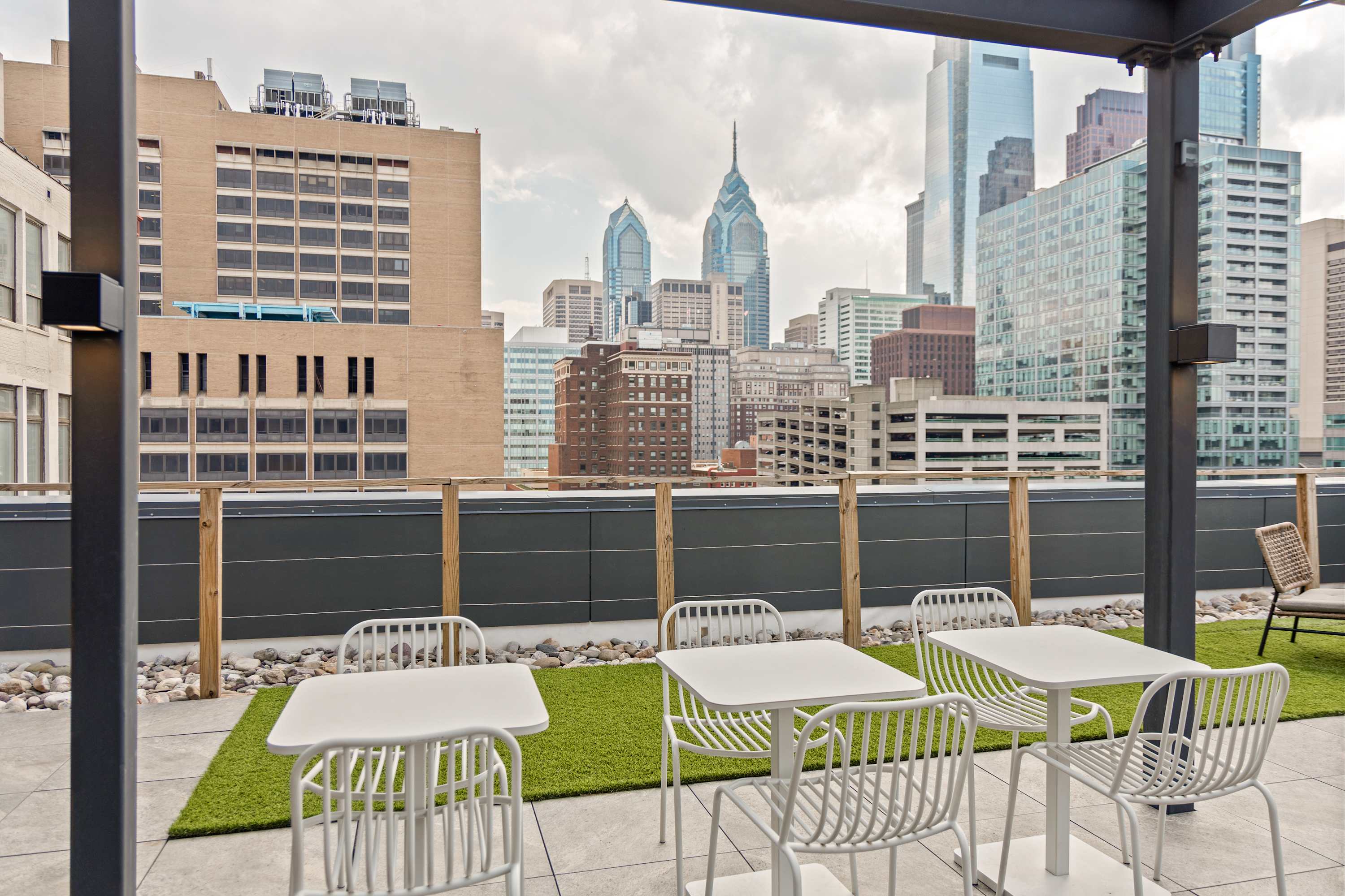 Rooftop deck with white outdoor tables and chairs set on artificial turf, offering panoramic views of a bustling city skyline with various high-rise buildings under a cloudy sky.