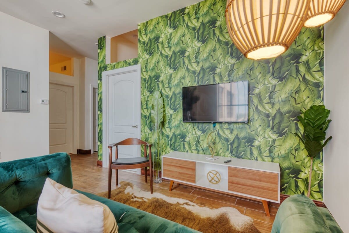 A cozy living space with a striking green banana leaf wallpaper accent wall, a wall-mounted TV above a mid-century modern media console, and a unique woven pendant light fixture.