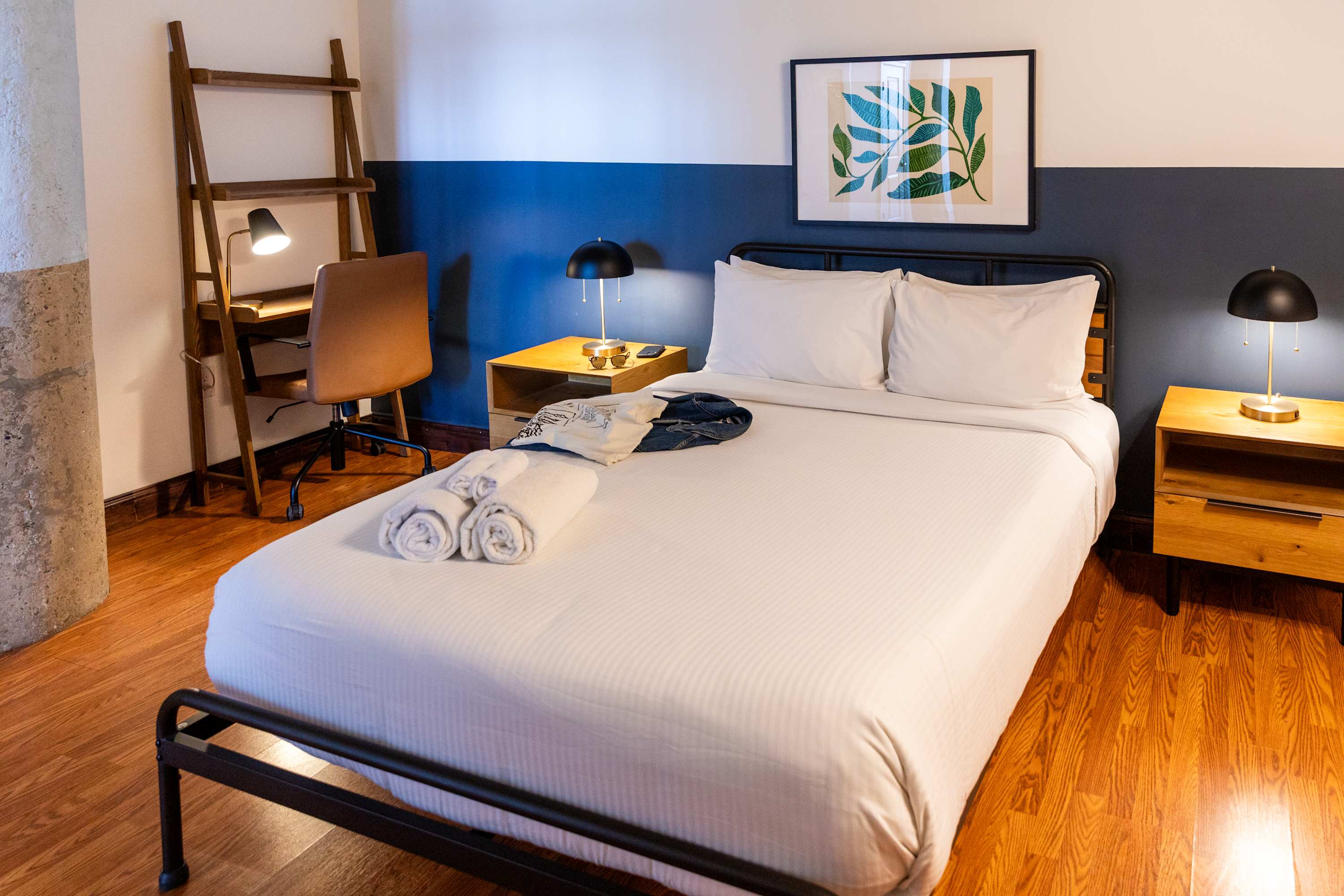 Cozy bedroom featuring a white bed with a black metal frame, two nightstands with lamps, and a desk with a brown office chair. The wall has a modern two-tone paint design in white and dark blue.