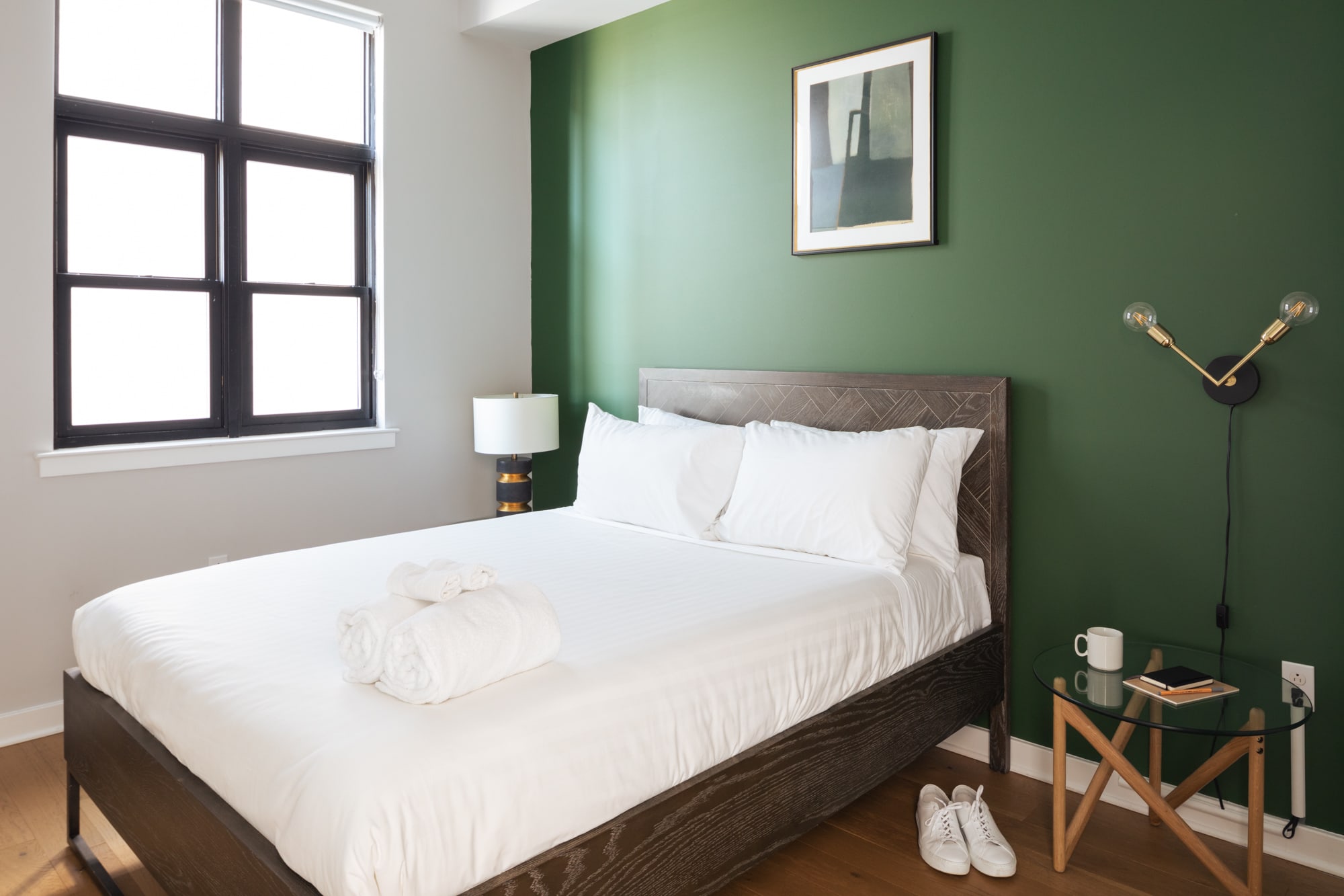 Cozy bedroom featuring a dark wood bed frame with white bedding and pillows, set against a vibrant dark green accent wall. A modern wall sconce and a glass-top nightstand with a mug and phone are visible on the right.