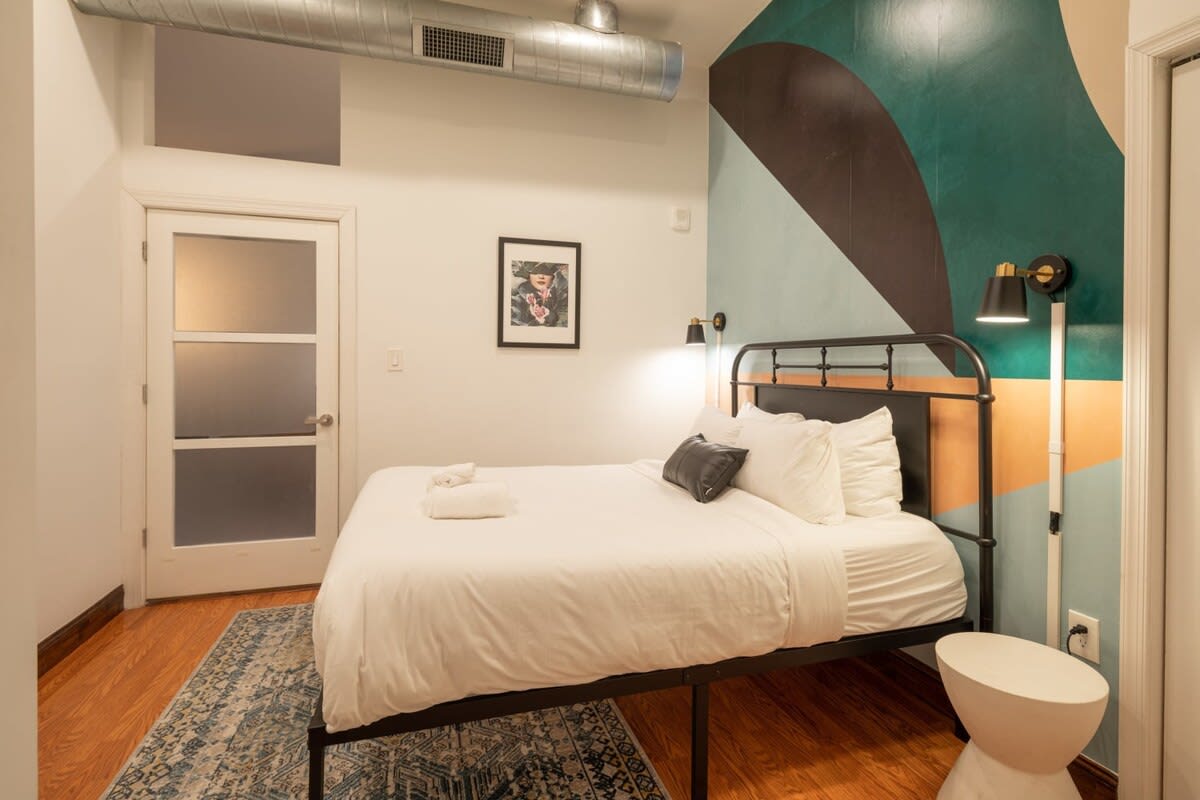 Cozy bedroom with a black metal bed frame, white bedding, and a striking geometric accent wall in teal, black, and orange, featuring a frosted glass door and a patterned area rug.