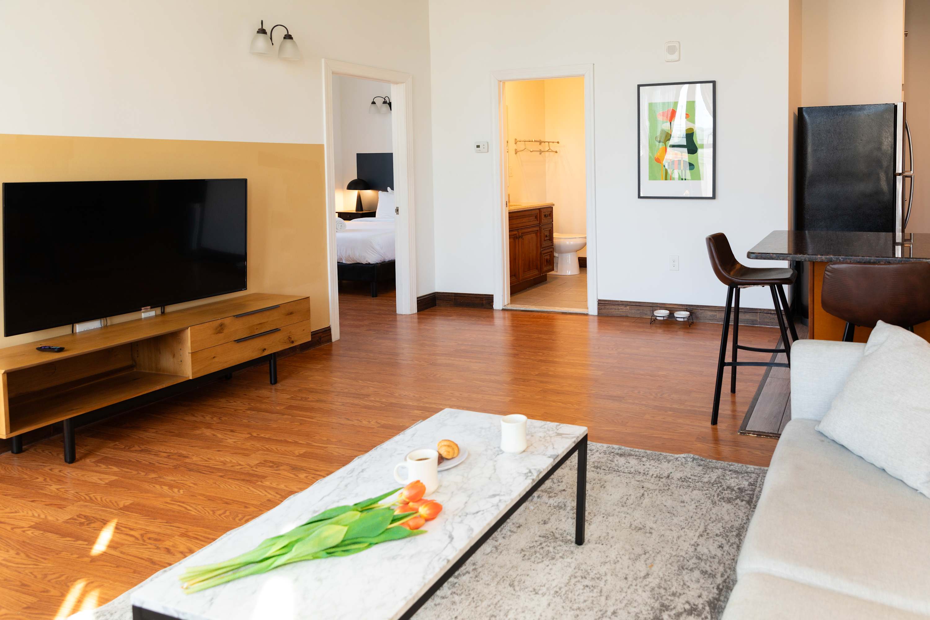 Open-concept living area with a flat-screen TV on a wooden console, a light sofa, and a marble coffee table. Hardwood floors extend towards a breakfast bar with stools and offer glimpses into a bedroom and bathroom through open doorways.