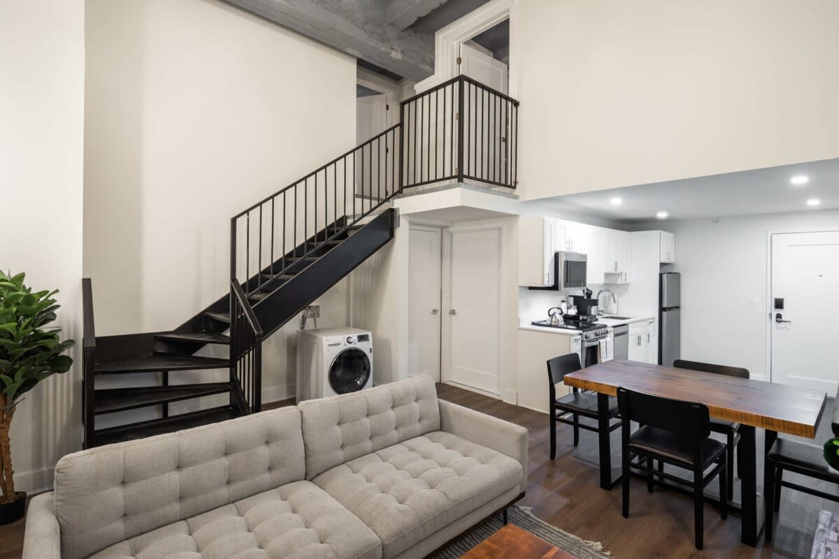 Open-concept living space showcasing a light gray sofa, a dining area, and a compact kitchen, with a striking black metal spiral staircase leading to an upper level and a washer/dryer unit below.