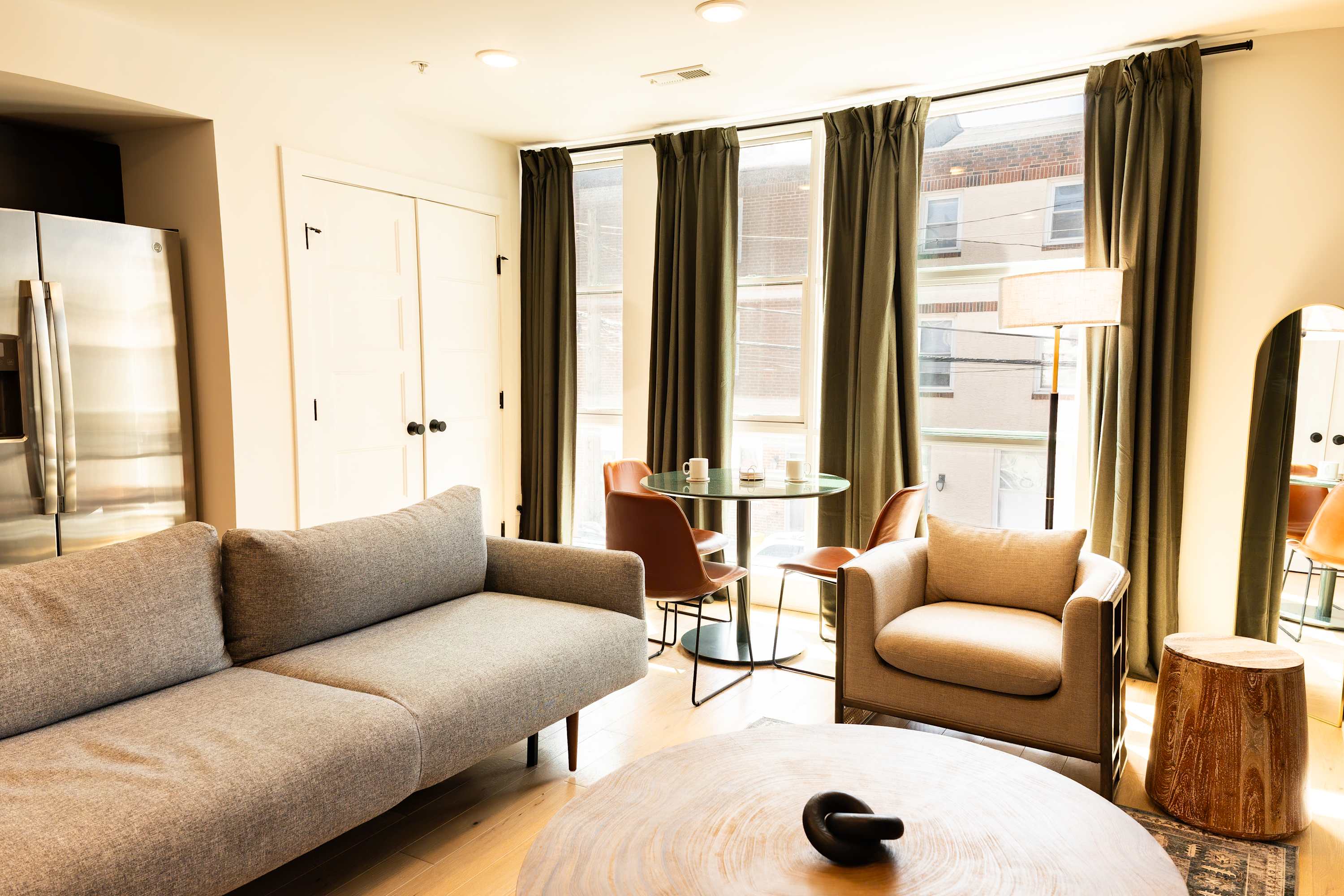 A bright living room with a light gray sofa and armchair, a round wooden coffee table, and a small dining area with a glass-top table and brown chairs by large windows with dark green curtains, all on light hardwood floors.