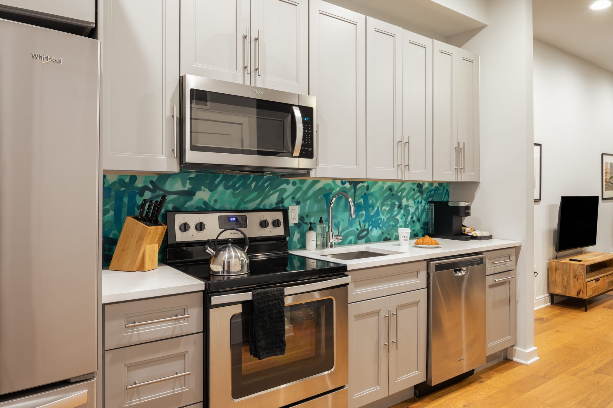 A modern kitchen featuring light grey shaker cabinets, stainless steel appliances including a microwave, stove, and dishwasher, and a vibrant teal and green abstract backsplash. White countertops and a stainless steel sink complete the functional space.