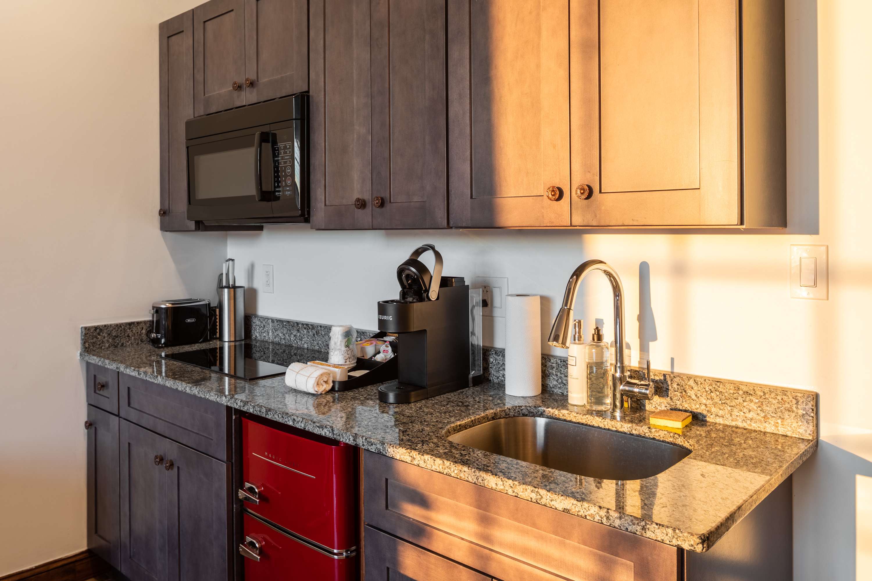 Compact kitchenette with dark gray shaker-style cabinets, speckled granite countertops, and a stainless steel sink. Appliances include a black microwave, a Keurig coffee maker, a toaster, and a retro red mini-fridge.