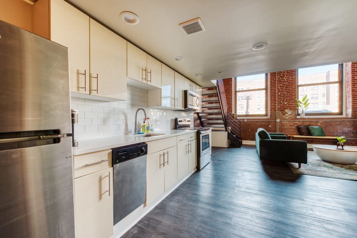 A modern kitchen with light beige flat-panel cabinets, white subway tile backsplash, and stainless steel appliances including a refrigerator, dishwasher, and microwave. The dark hardwood floors extend into the open living area visible in the background.
