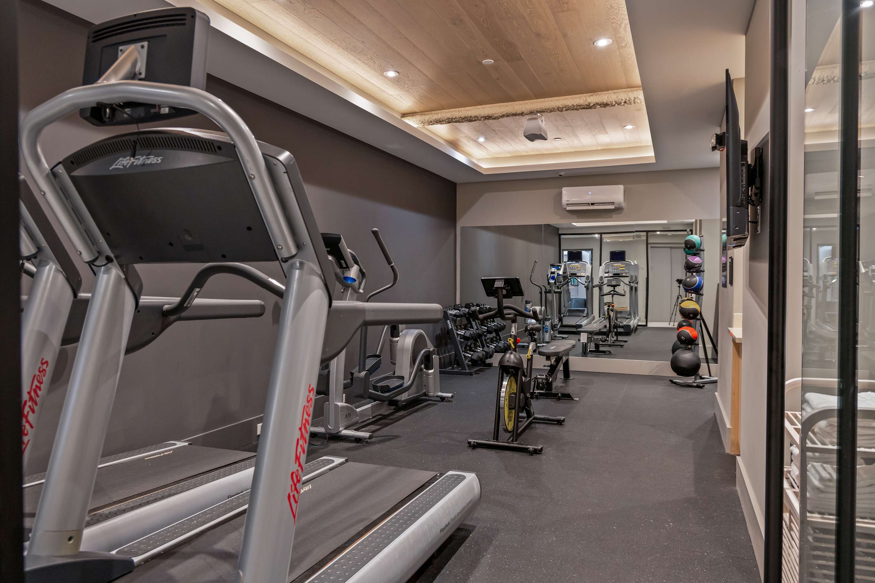 Well-equipped fitness center featuring treadmills, an elliptical machine, free weights, and an exercise bike, all set against dark gray walls with a large mirror reflecting the equipment. A unique wooden ceiling panel with recessed lighting adds warmth to the space.