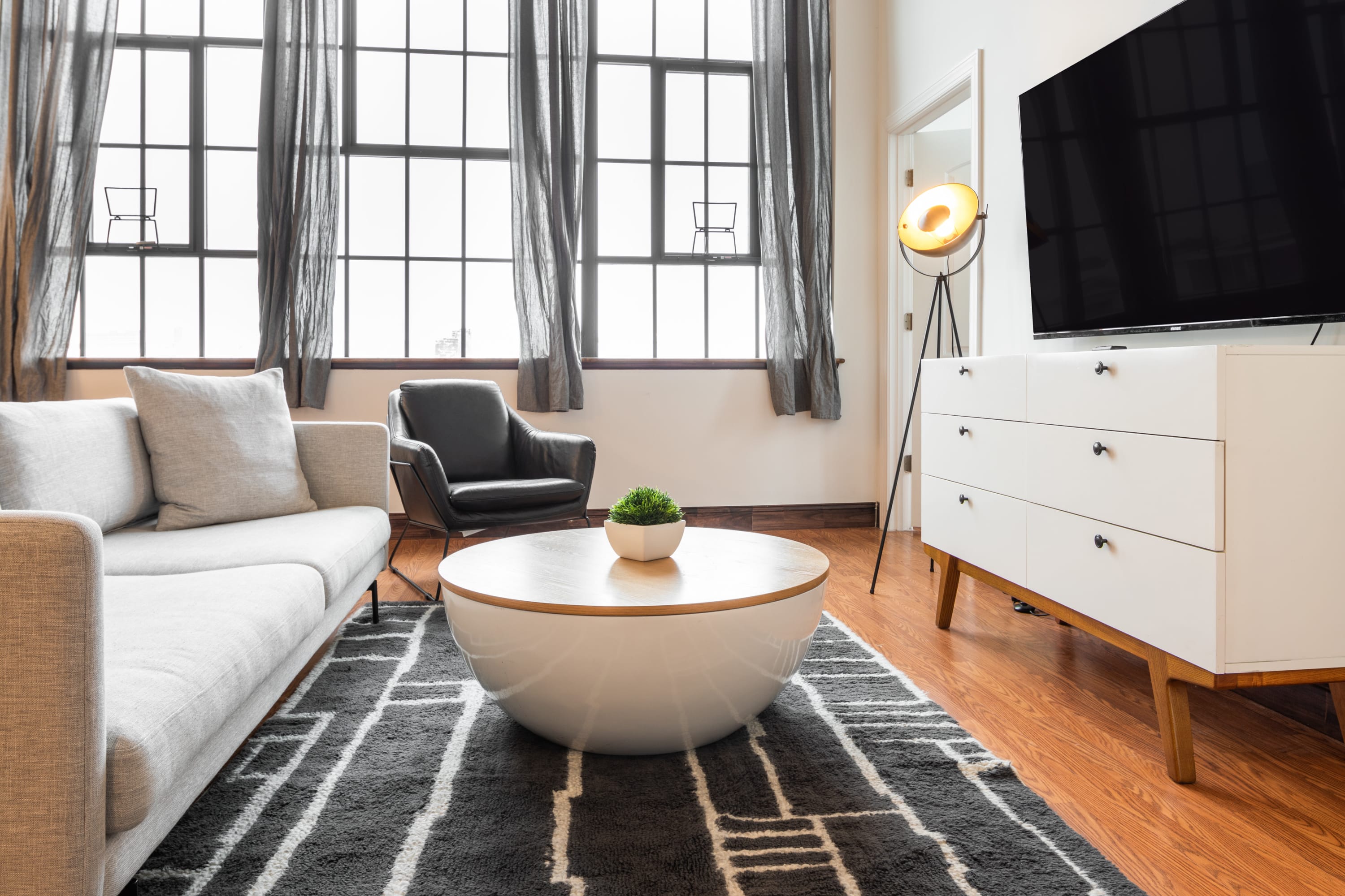 Bright living room featuring a light gray sofa, a black leather armchair, and a round white coffee table with a wooden top on a geometric patterned rug. Large industrial windows with gray curtains illuminate the space, and a TV is mounted above a white dresser.